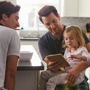 A man in pyjamas with a young girl also in pyjamas sitting on his knee whilst they are both looking at a tablet he is holding and another man is sitting with them.